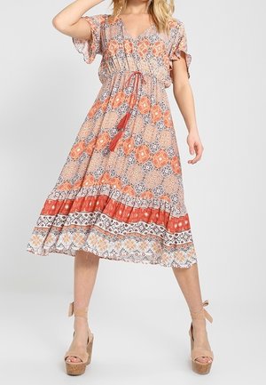 Kvinde i en knælange, mønstret kjole med orange og beige nuancer, iført beige platformhøjhælede sko, poserer med den ene arm bøjet og benene spredt.