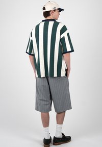 Joven vestido con una camisa de manga corta con rayas verticales blancas y verde oscuro, pantalones cortos gris a rayas finas, calcetines blancos, zapatos negros y una gorra blanca.
