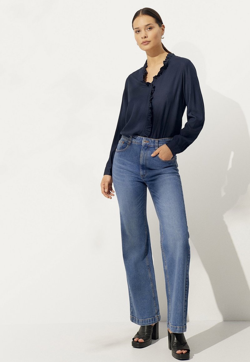 Navy blouse met ruchekraag, gecombineerd met hoge blauwe spijkerbroeken. Model draagt zwarte platformhakken en staat tegen een lichte achtergrond.