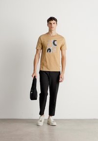Beige bomull T-skjorte med abstrakt grafisk trykk, parret med svarte tapered bukser og hvite sneakers. Modellen holder en svart veske.