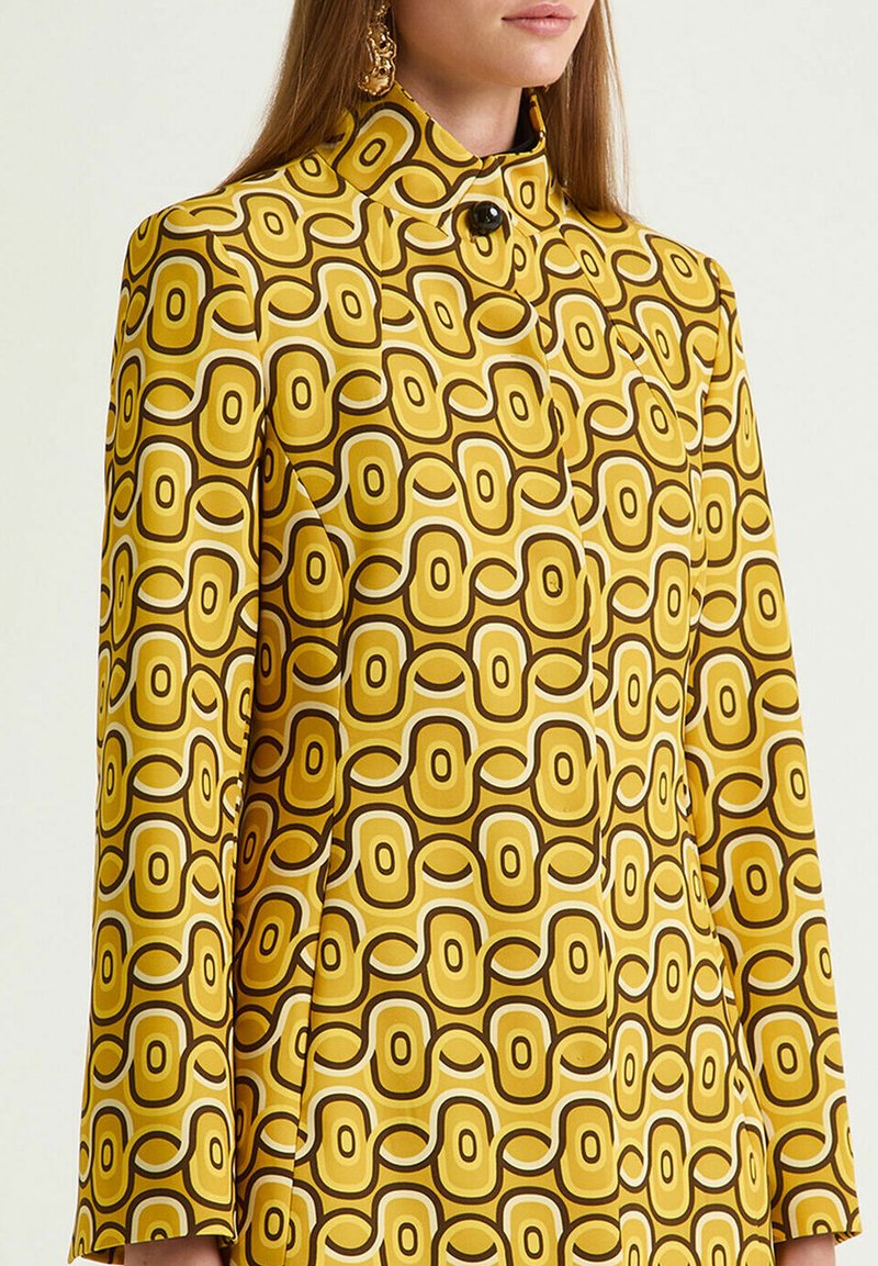 Veste à manches longues en tissu jaune vif présentant un motif géométrique marron et blanc, col montant et fermeture à boutons noirs.