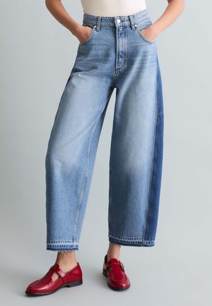 Osoba nosząca szerokie, dwukolorowe niebieskie dżinsy z surowymi wykończeniami i czerwone skórzane buty na klamrę, stojąca z rękami w kieszeniach.