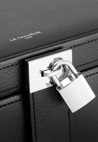 Bolso de cuero negro con un cierre y un candado plateados. Presenta una superficie texturizada y costuras visibles. Logo "Le Tanneur 1898" grabado en la parte superior.