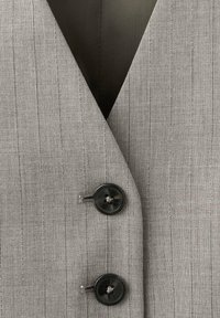 Chaleco gris a rayas con un escote en V profundo, que presenta dos botones negros y una tela texturizada. Forro interior visible en el cuello.