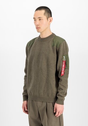 Man draagt een olijfgroene trui met schouderstukken en een ritssluiting zak op de arm met een rood "Remove Before Flight" label.