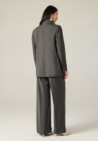Completo grigio con giacca su misura e pantaloni a gamba larga, realizzato in tessuto testurizzato. La giacca presenta una singola spacchetta posteriore e dettagli con bottoni.