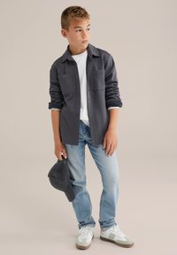 Pojke i mörkgrå knappjacka över vit skjorta, ljusblå jeans, vita sneakers, håller en mörkgrå keps, tittar åt sidan.