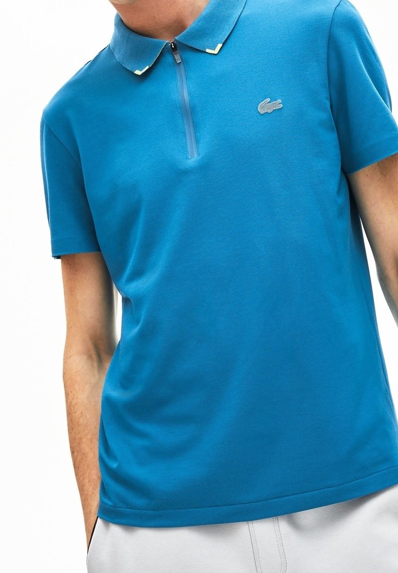 Homme portant un polo bleu à manches courtes avec col zippé et petit logo crocodile sur la poitrine gauche.