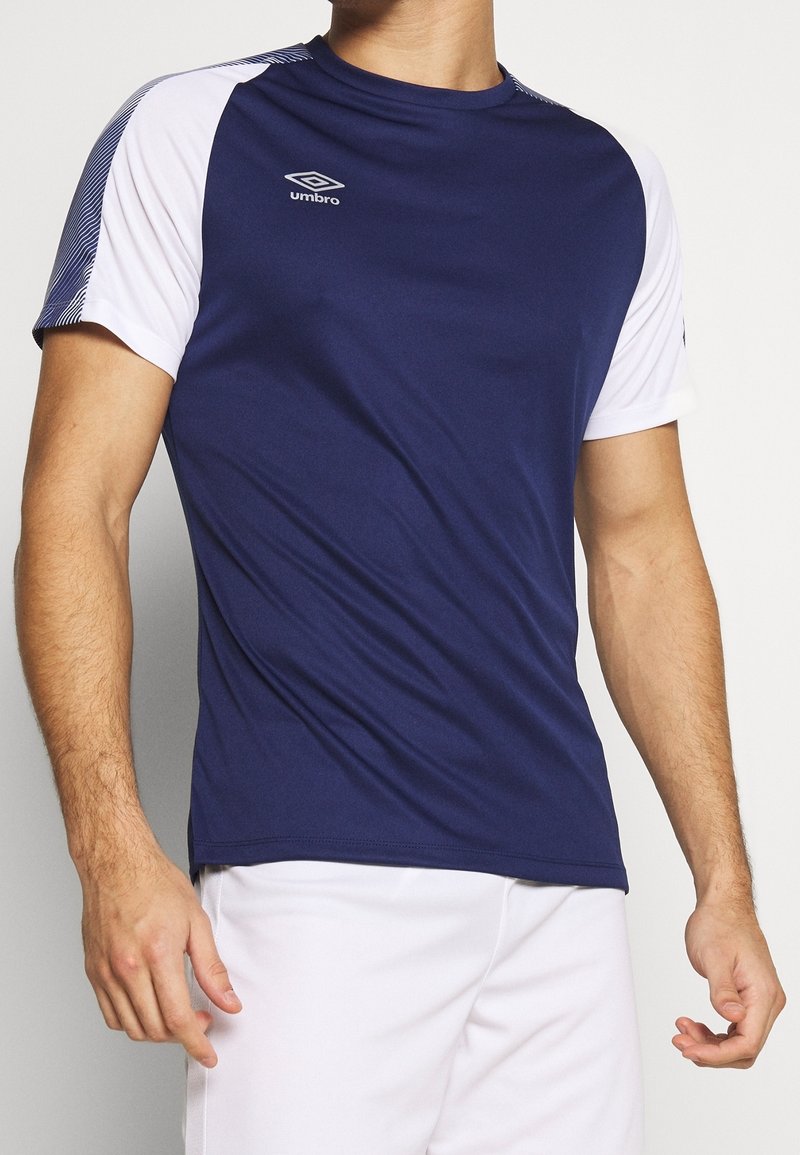 Námořnická modrá sportovní košile s bílými krátkými rukávy, se pruhovaným vzorem na rameni a logem na hrudi. Vyrobena z lehkého materiálu.