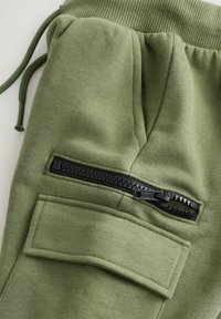 Groene katoenen joggers met een ribbelrand aan de tailleband, een verstelbaar trekkoord en een zwarte ritszak met daarboven een rechthoekige patch.