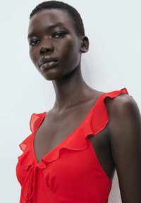 Robe rouge à volants avec bretelles spaghetti, présentant un détail à nouer à l'avant. Tissu lisse avec une légère texture, corsage ajusté.