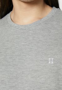 Primo piano di una persona che indossa un maglione grigio chiaro a collo rotondo con un piccolo "II" ricamato sul petto.