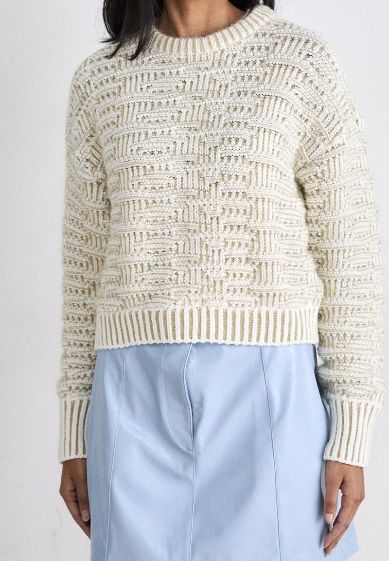 Femme portant un pull en maille crème avec un motif géométrique texturé et une jupe en cuir bleu clair, vue du cou aux hanches.