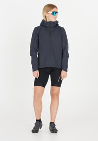 Donkerblauwe lichte jas met een capuchon, een ritssluiting aan de voorkant en reflecterende details, gecombineerd met zwarte fietsshorts en zwarte sportschoenen.