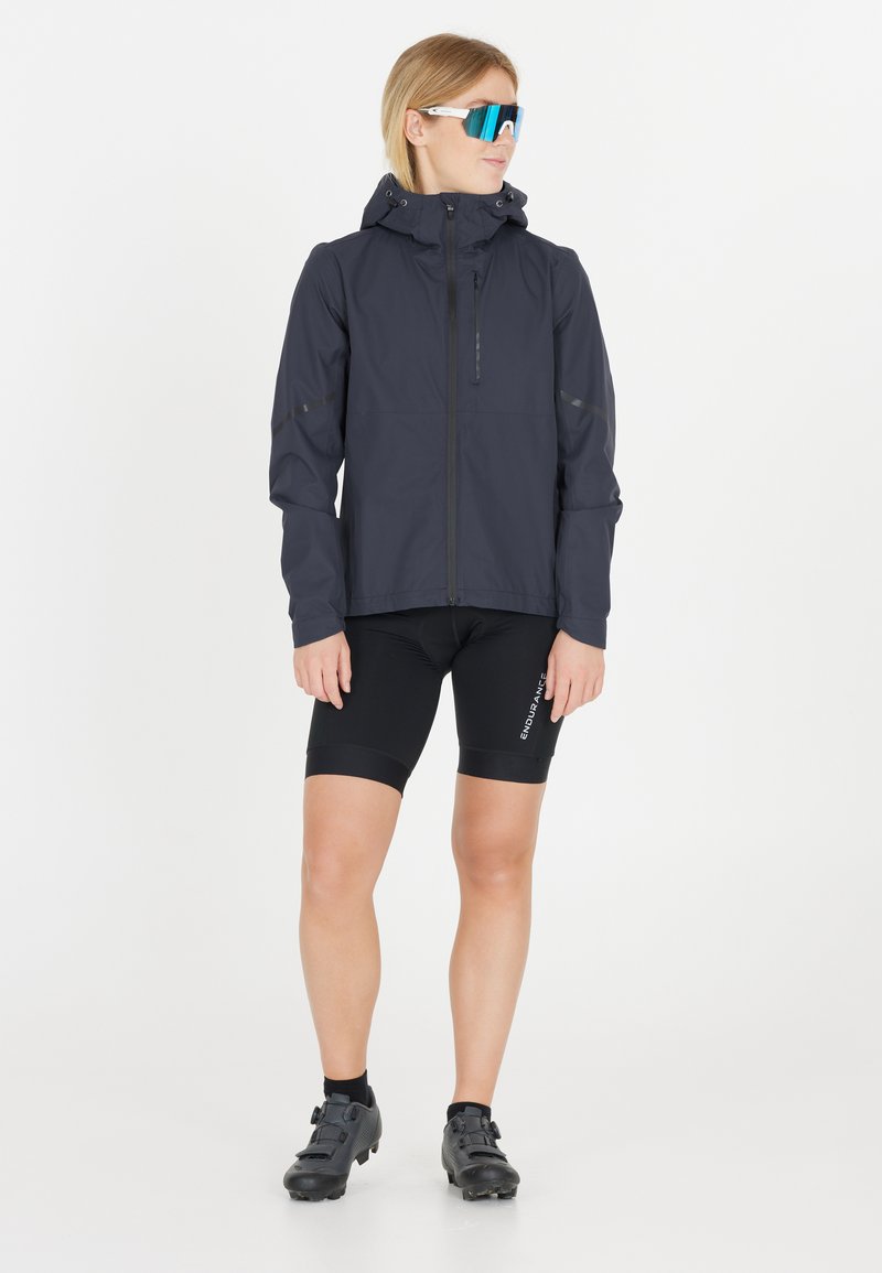 Donkerblauwe lichte jas met een capuchon, een ritssluiting aan de voorkant en reflecterende details, gecombineerd met zwarte fietsshorts en zwarte sportschoenen.