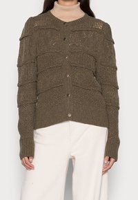 Cardigan vert olive avec des rayures texturées, fermeture à boutons et poignets côtelés. Porté sur un col roulé beige, il présente un design en tricot confortable.