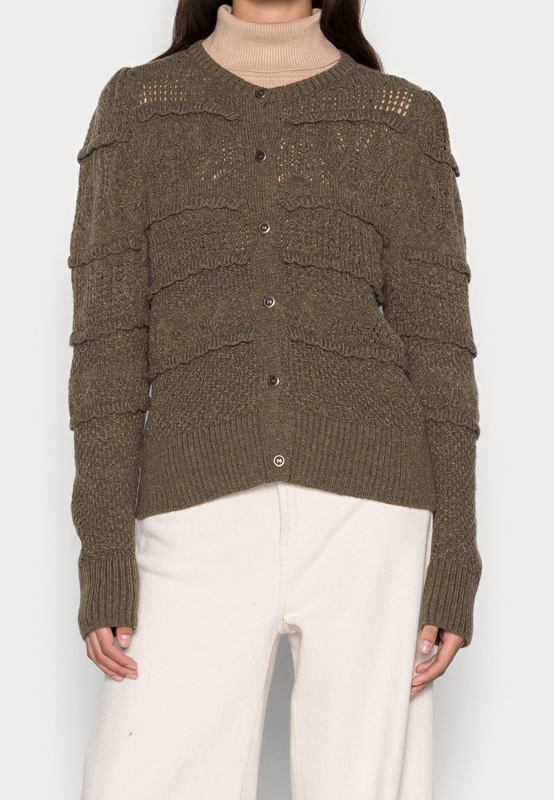 Cardigan vert olive avec des rayures texturées, fermeture à boutons et poignets côtelés. Porté sur un col roulé beige, il présente un design en tricot confortable.