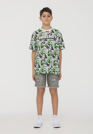 T-shirt grafico presenta un design di Minecraft in nero e verde con personaggi e loghi pixelati. Indossato con pantaloni corti in denim grigi e sneaker bianche.