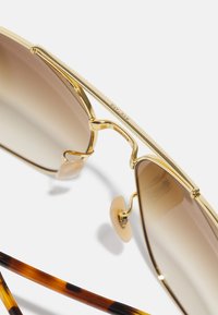 Ochelari de soare din metal auriu, cu lentile de culoare maroGradient, cu suporturi pentru nas transparente și brațe cu model țestoasă, având un design elegant și unghiular.