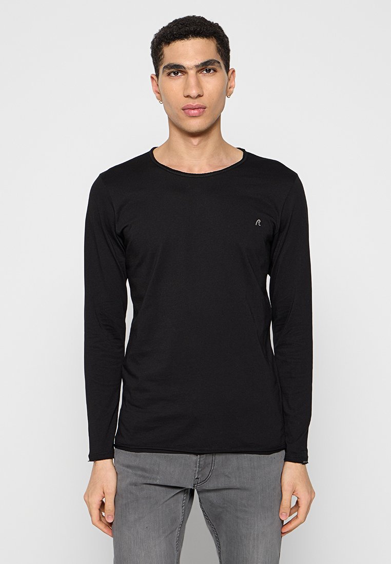 Junger Mann in einem eng anliegenden schwarzen Langarmshirt und grauen Jeans, der vor einem schlichten weißen Hintergrund steht.