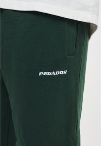 Groene katoenen joggers met zijzakken, voorzien van een witte geborduurde "PEGADOR" logo op de linkerdij. Glad materiaal, casual pasvorm.