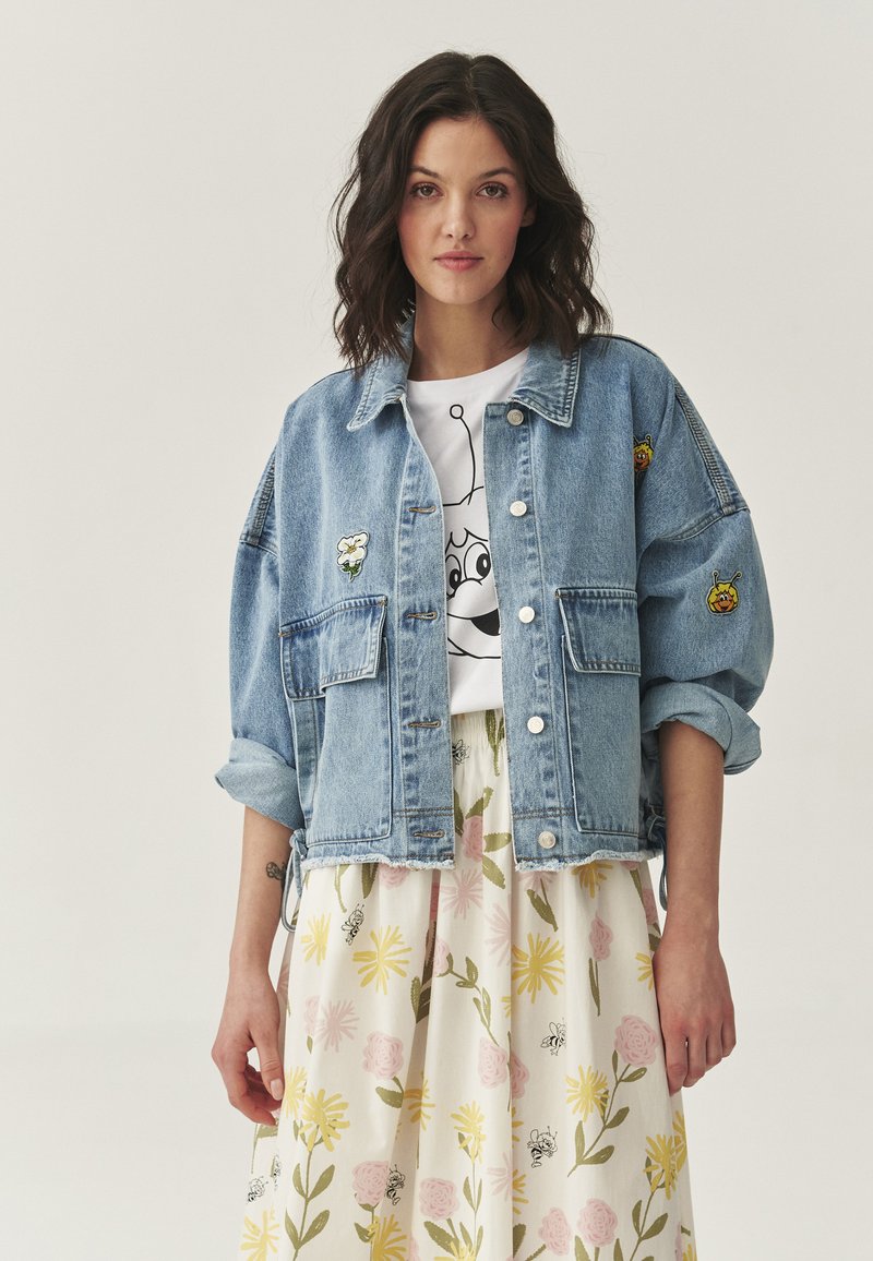 Vrouw met donker golvend haar in een spijkerjack met patches, een wit T-shirt met opdruk en een witte rok met gele en roze bloemenprints.