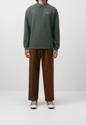 Osoba ubrana w ciemnozieloną koszulkę polo z długim rękawem, brązowe spodnie o szerokich nogawkach oraz ciemnobrązowe buty sportowe z białymi podeszwami, stojąca na jednolitym tle.