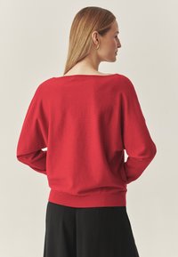 Pull rouge, coupe ample, col rond, poignets côtelés, texture douce, avec une vue arrière montrant une longueur légèrement plus courte. Stylisé avec un pantalon noir.