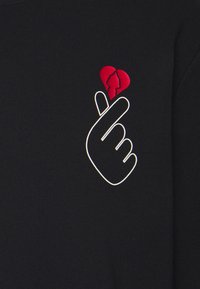 Sudadera negra con un contorno blanco de una mano haciendo un gesto de corazón, con un pequeño detalle en forma de corazón rojo.