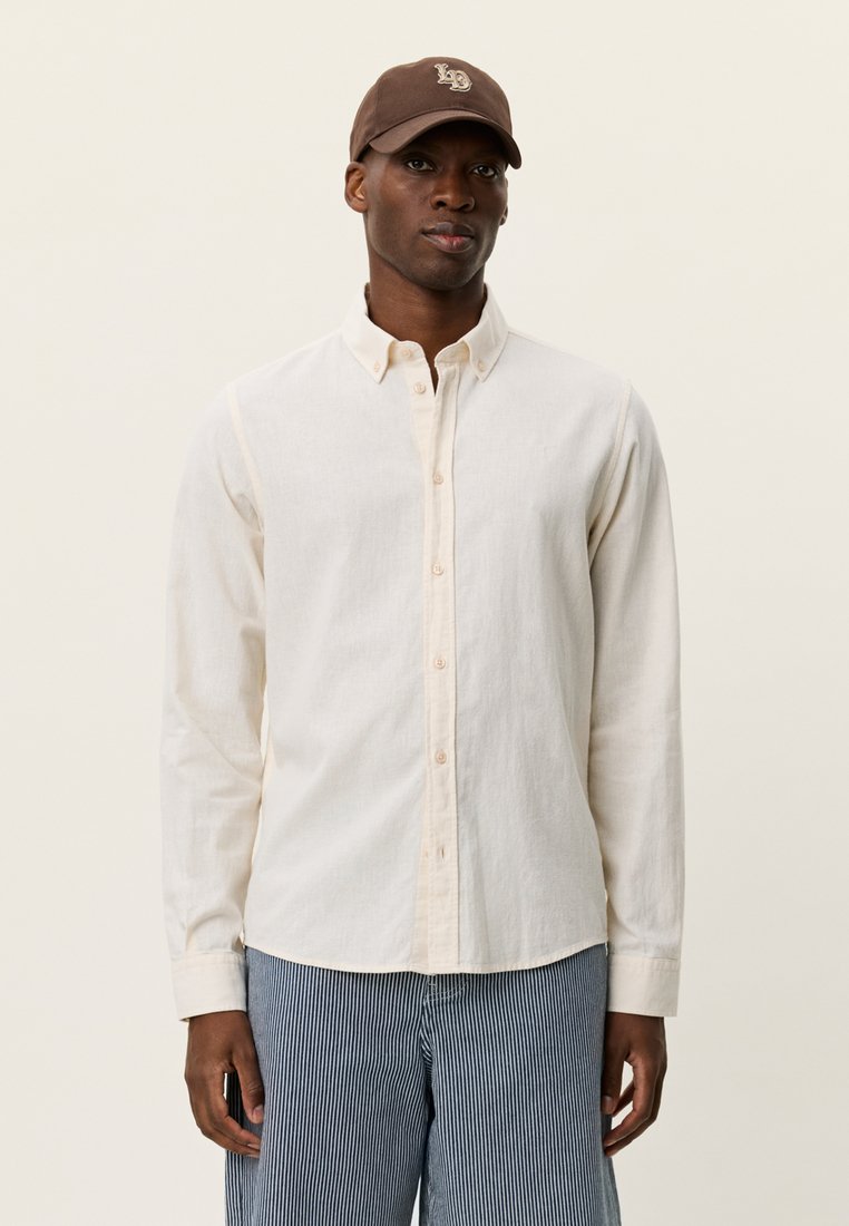 Φωτεινό μπεζ πουκάμισο με κουμπιά από βαμβάκι, με μυτερό γιακά, μακριά μανίκια και απαλή υφή, συνδυασμένο με ριγέ μπλε παντελόνια και καφέ καπέλο.