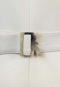 Tela blanca con una sutil textura diagonal que presenta una hebilla cuadrada y translúcida grabada con "PATRIZIA PEPE" y una letra 'P' estilizada.