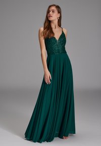 Femme portant une robe de soirée sans manches vert foncé avec un corsage pailleté et une jupe fluide, debout devant un fond gris uni.