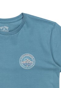 Camiseta de algodón azul con cuello redondo, que presenta un logo circular en colores pastel que representa montañas y un sol, con el texto "Billabong Adventure Division".