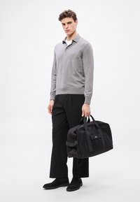 Geantă de duffle din țesătură neagră cu mânere scurte combinate și curea de umăr. Are închidere cu fermoar și detaliu cu logo pe față.