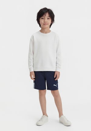 Chlapec stojaci v bielej košeli s dlhým rukávom, námorníckych modrých šortkách Puma a bielych teniskách na jednoduchom bielom pozadí.