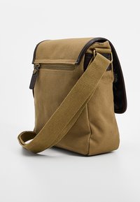 Ruskea kangaslaukku olkapäällä kannettavaksi, jossa on vetoketjullinen tasku, nahkaiset yksityiskohdat, säädettävä remmi ja jämäkkä suorakaiteen muotoinen rakenne. Pinnan teksturoitu yksityiskohta.