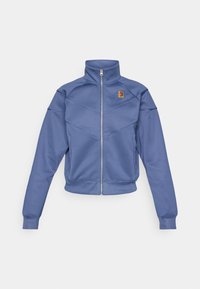 Chaqueta azul con cremallera y collar alto, presenta un diseño geométrico y un pequeño logo en el lado izquierdo del pecho. Está hecha de una tela suave.