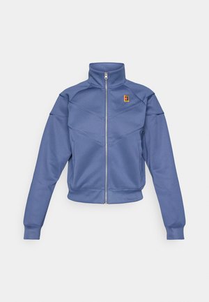 Blauwe zip-up jas met een hoge kraag, met een geometrisch ontwerp en een klein logo op de linkerborst. Gemaakt van een gladde stof.