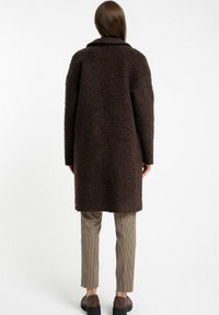 Brązowy, puszysty płaszcz z kołnierzem i długimi rękawami, o luźnym kroju i fakturowanym materiale. Nosi się go na dopasowane, w kratę spodnie.