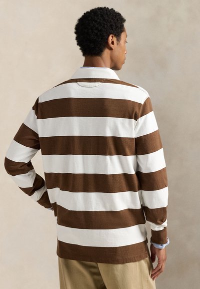 Uomo con le spalle rivolte verso di noi, che indossa una camicia a righe orizzontali marroni e bianche a maniche lunghe e pantaloni beige su uno sfondo beige uniforme.