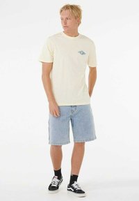 Kortærmet lys gul t-shirt med et blåt logo på brystet, parret med lyseblå denimshorts og sorte sneakers.