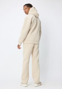 Sweat à capuche en polaire beige avec poche kangourou et poignets côtelés, associé à un pantalon de jogging beige assorti. Le mannequin est debout, légèrement tourné vers l'arrière, portant des baskets.