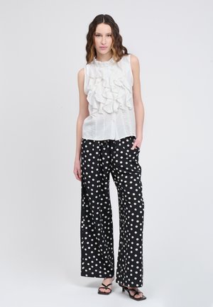 Weißes ärmelloses Oberteil mit geschichteten Rüschen; kombiniert mit weitgeschnittenen schwarzen Hosen mit großen weißen Polka-Dots und offenen schwarzen High Heels.