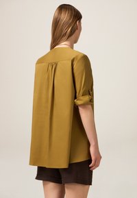 Senfgelbe Bluse mit lockerer Passform, gefaltetem Rückendetail und aufgerollten Ärmeln. Leichter Stoff und gerader Saum.