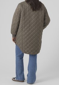 Cappotto imbottito verde oliva dal design lungo, con polsini a coste e spacchi laterali, abbinato a jeans blu svasati e scarpe da ginnastica nere.