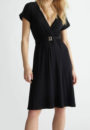 Kobieta ubrana w czarną sukienkę o długości do kolan, z krótkimi rękawami i czarnym paskiem z złotą klamrą w talii.