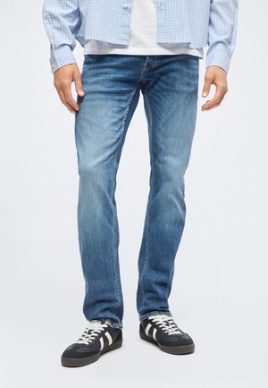 Underkroppen af en person iført lyseblå jeans, sorte og hvide sneakers, en hvid skjorte og en blå ternet button-up skjorte.
