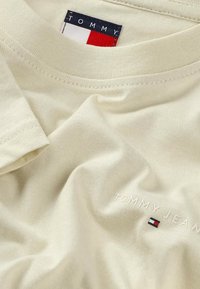 Camiseta Tommy Jeans color crema que muestra el cuello, la textura de la tela y el logo bordado con detalle de bandera en rojo, blanco y azul.