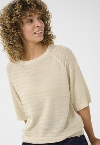 Pull en tricot beige à manches courtes avec un motif rayé texturé, doté d'un col rond et d'un ourlet côtelé, porté par-dessus un pantalon sombre.