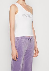 Bílý top na jedno rameno s přiléhavým střihem a grafickým potiskem "JUICE" vyloženým z kamínků, doplněný o světle fialové velurové kalhoty se zipovými kapsami.